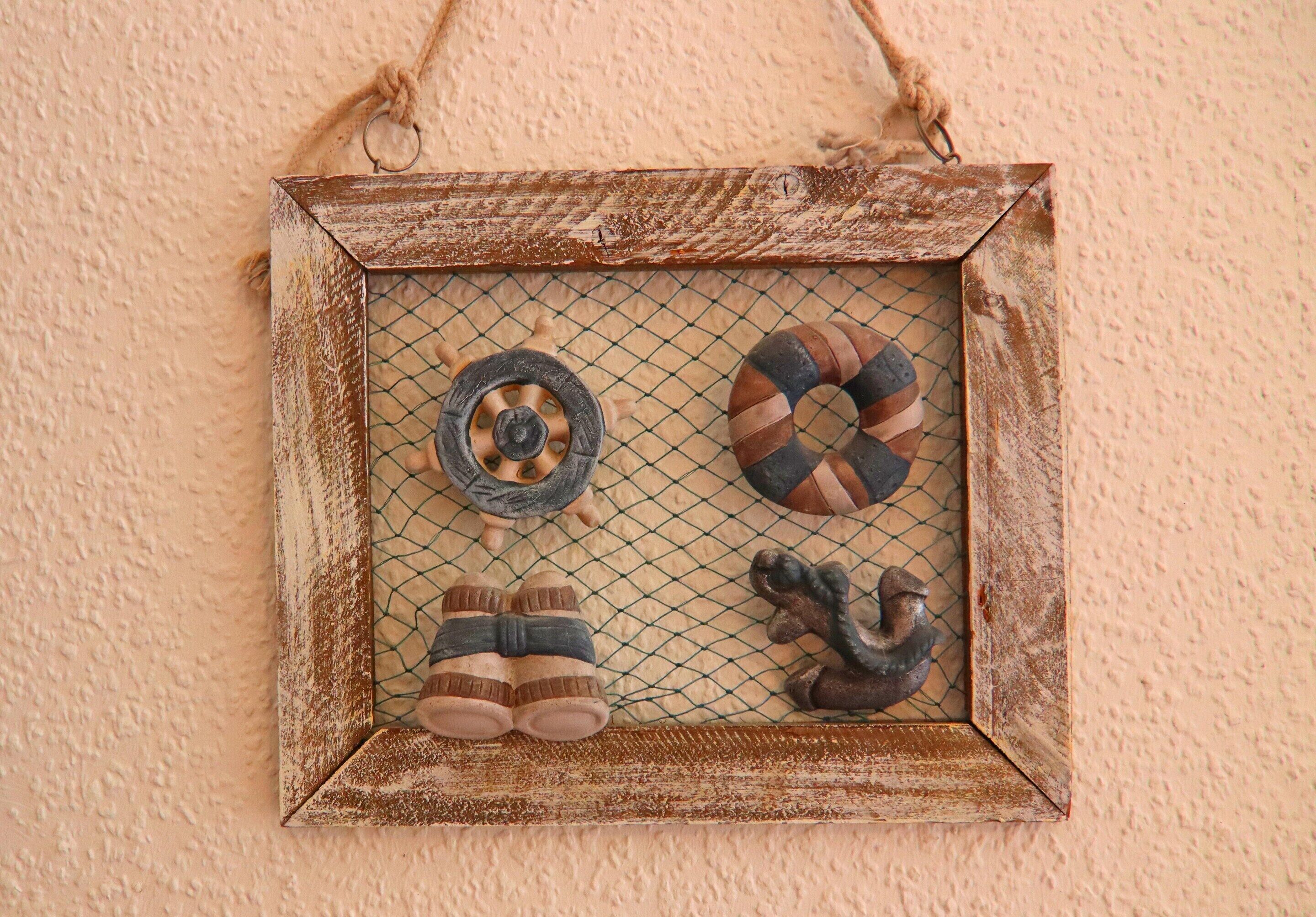 A rustic, weathered metal frame with a rope handle, containing a netted display of a tire, a life preserver, a pair of boots, and a small figure.