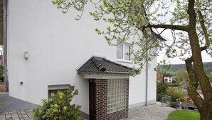 Un bâtiment avec une petite extension en briques, une cour pavée et un arbre aux fleurs jaunes.