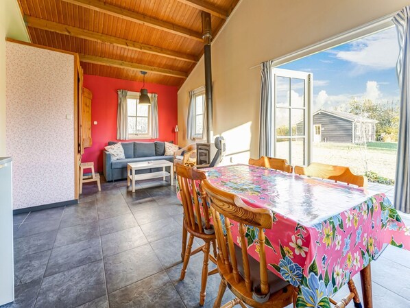 A dining area with a table covered in a floral tablecloth, wooden chairs, and a view of the outdoors through large windows.