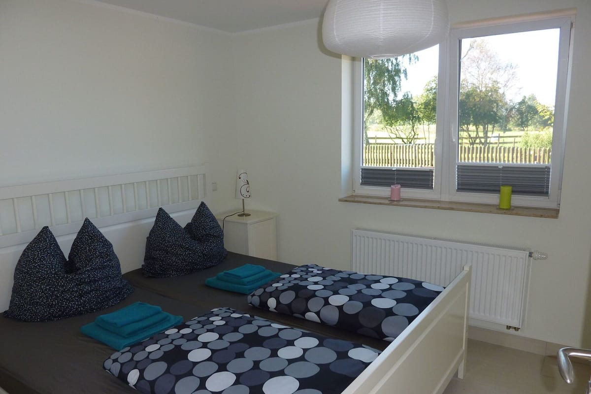 Une chambre avec deux lits, une fenêtre avec des stores, un radiateur et une petite table avec une lampe.