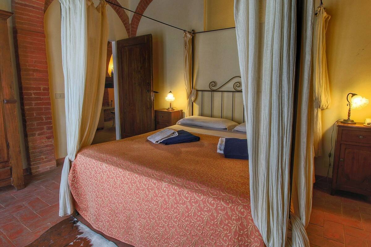 Un lit à baldaquin avec une couvre-lit à motifs rouges, une table de chevet en bois avec une lampe, et un mur de briques avec une porte en arc de cercle.