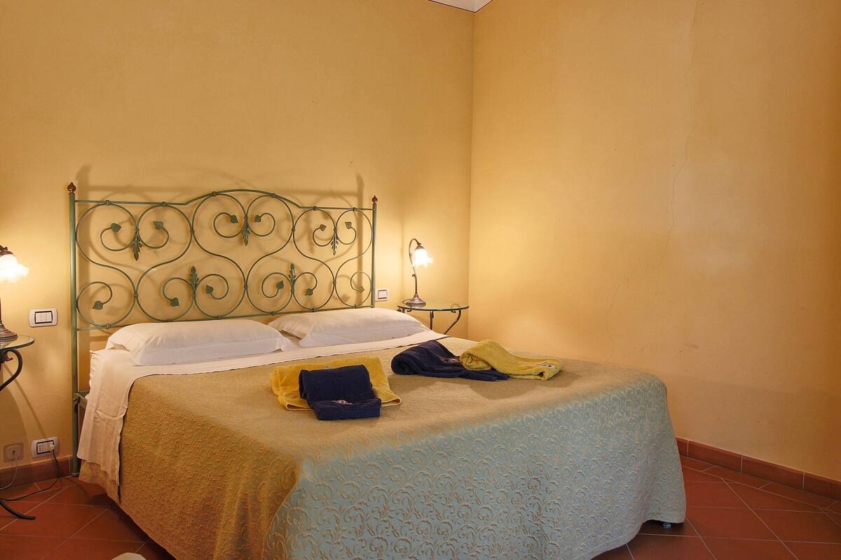 Un lit simple avec une tête de lit en métal, deux tables de chevet avec des lampes et une couvre-lit à motifs.