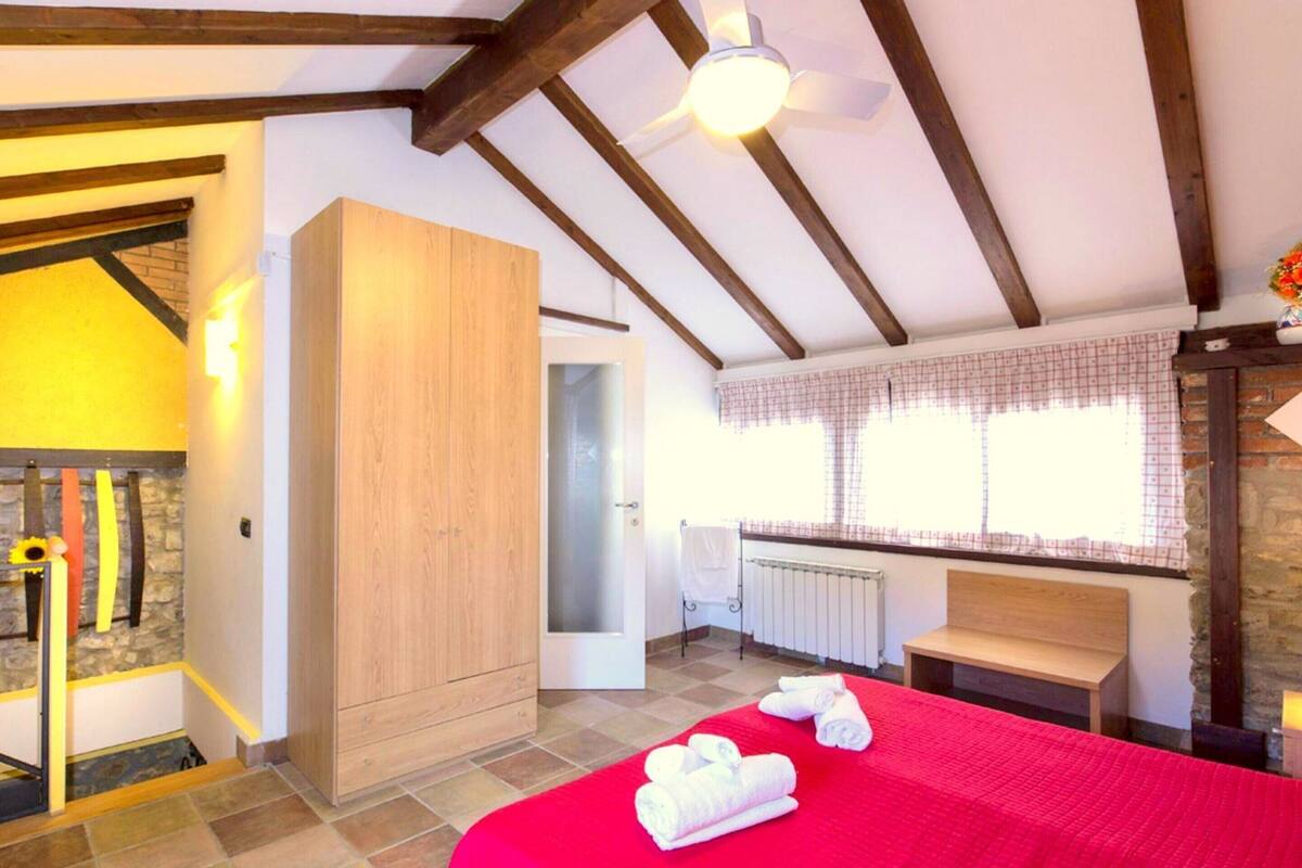 A bedroom with a red bedspread, wooden wardrobe, and a ceiling fan.