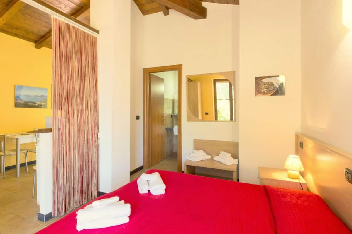 A hotel room with a red bedspread, a wooden ceiling, a desk, a chair, a bathroom with a mirror, and towels on the bed.