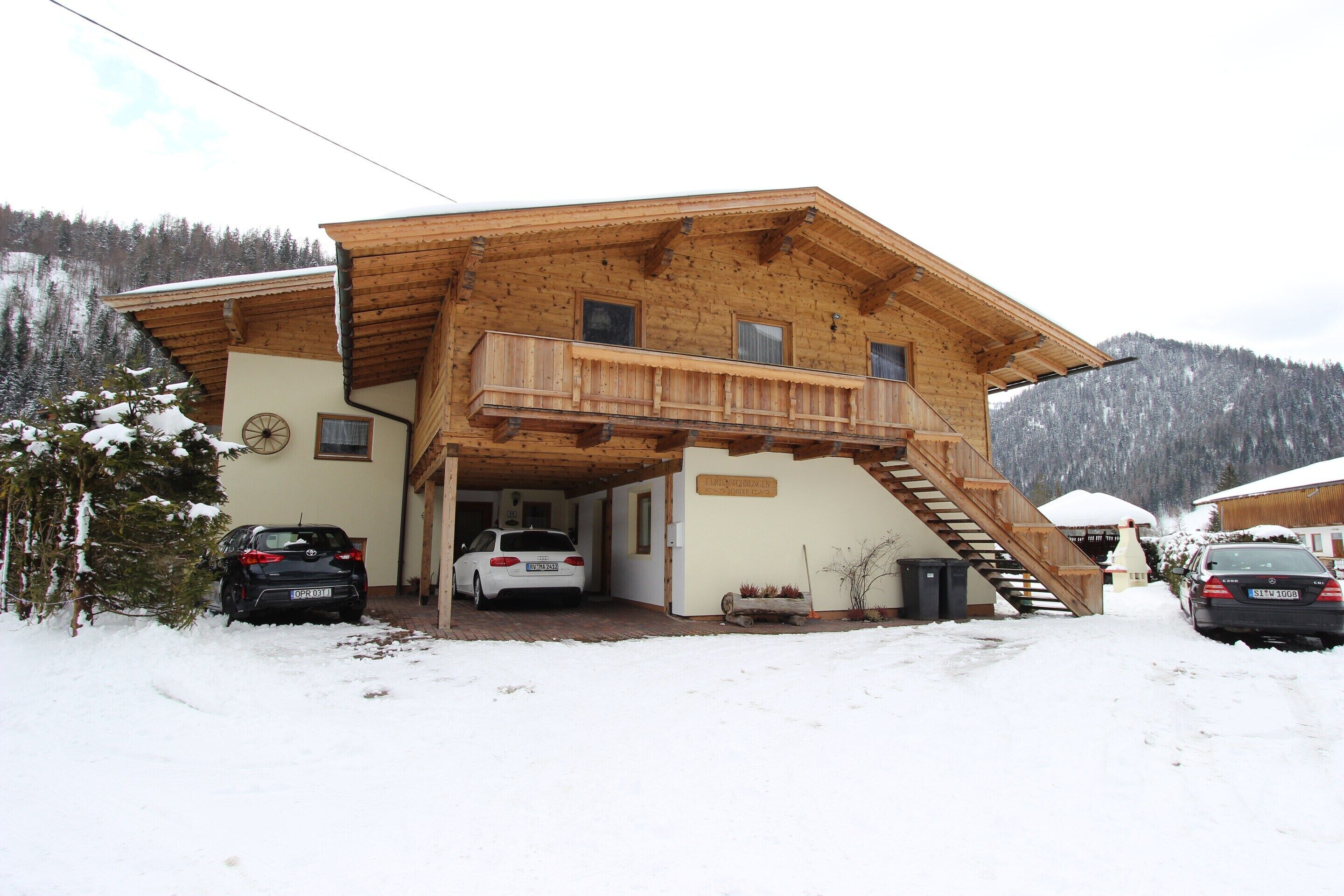 Eine Holzhütte mit Balkon, zwei Autos davor und ein verschneiter Berggipfel im Hintergrund.