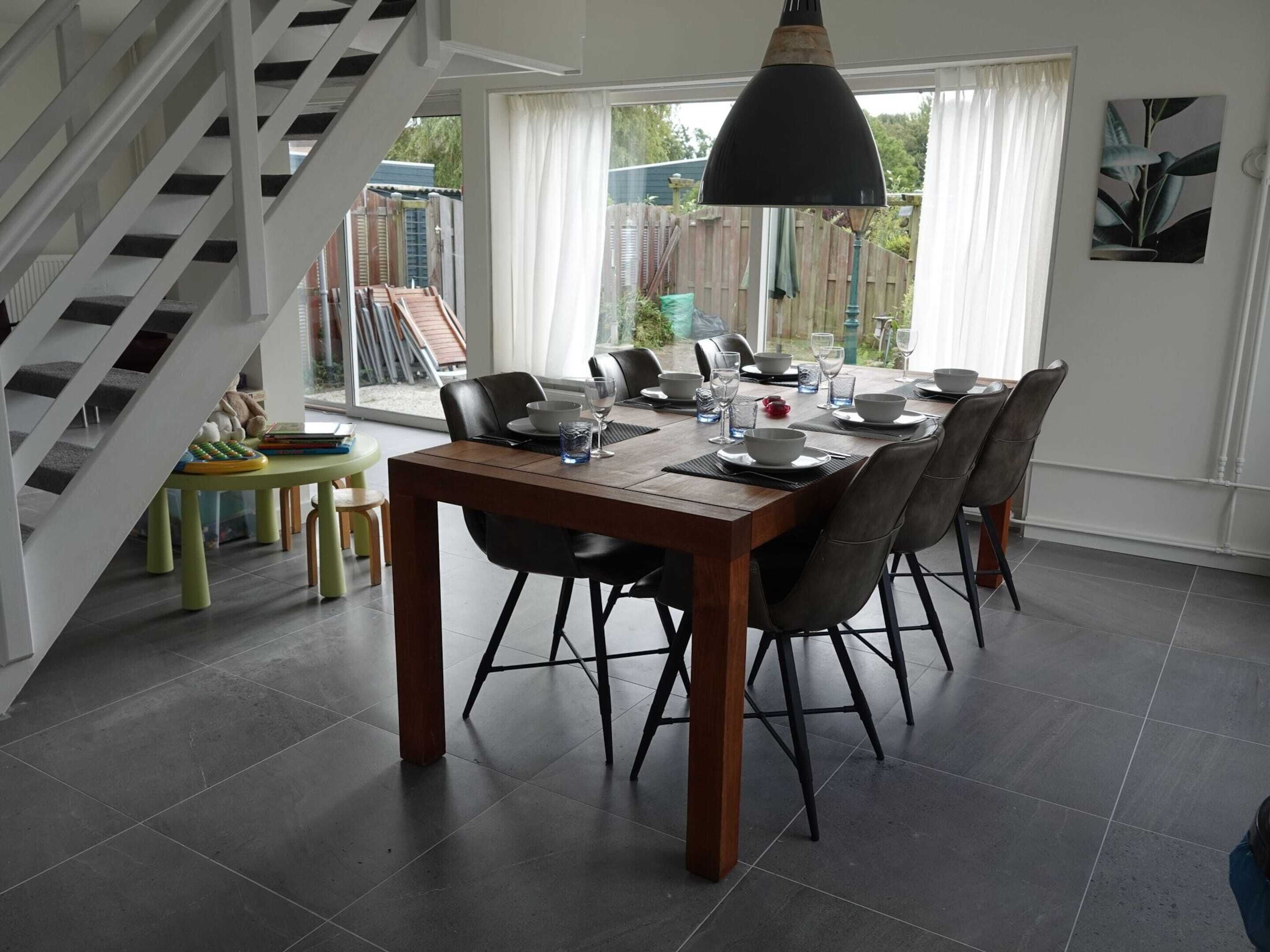 A dining area with a wooden table set for six, surrounded by chairs, a pendant light, and a view of the outdoors.
