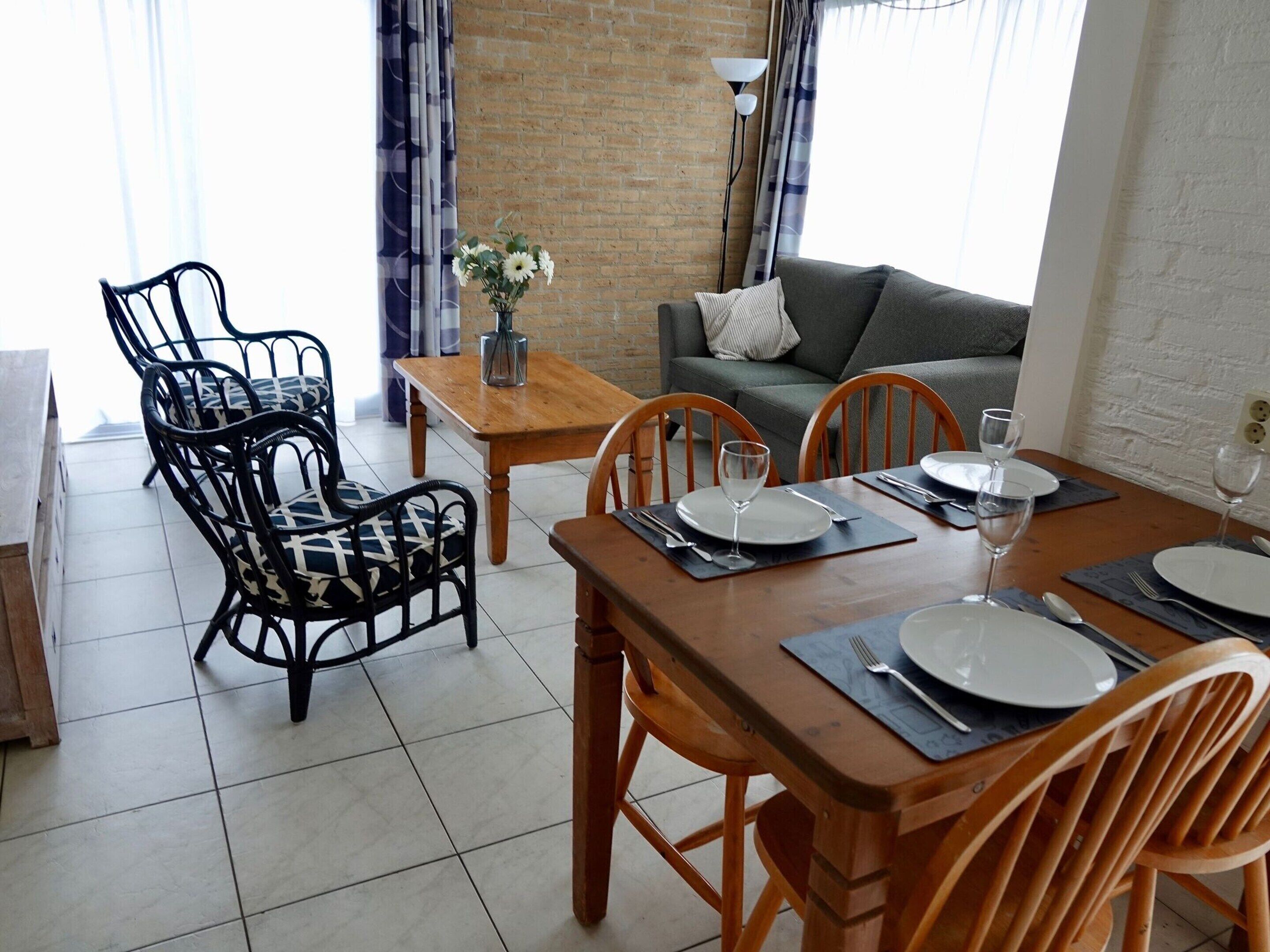 Un comedor con una mesa de madera dispuesta para cuatro personas, rodeada de sillas de metal negro. En el fondo, un sofá gris y una pared de ladrillo.