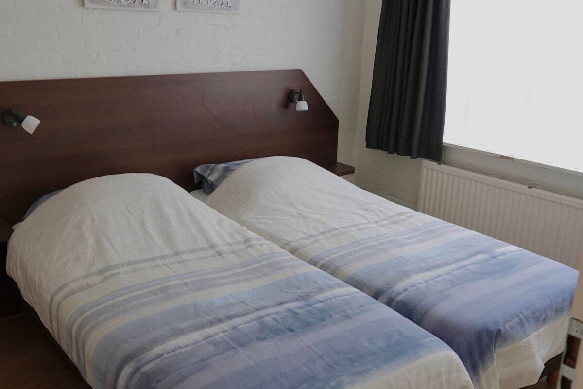Ein Doppelbett mit gestreifter Bettdecke in einem Zimmer mit Holzkopfstütze, Nachttisch und einem Vorhangfenster.