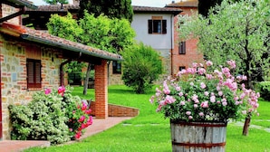 Un bâtiment rustique en pierre, doté d’une cheminée en briques, entouré d’une végétation luxuriante et de buissons en fleurs.