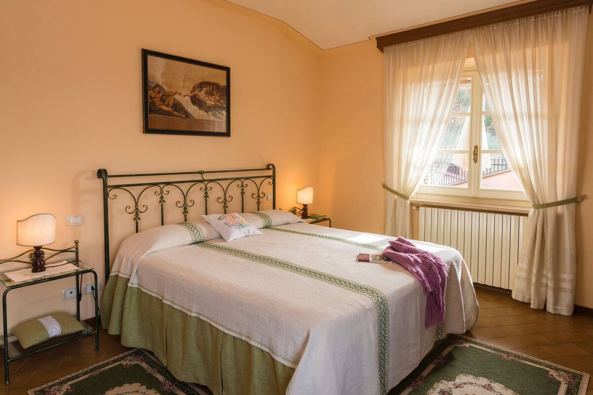 A bedroom with a bed, a nightstand, a lamp, a window with curtains, and a painting on the wall.