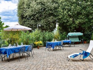 An outdoor patio with tables and chairs, a striped umbrella, and a large tree providing shade. - Air-conditioned Flat With Pool and Wellness Area (Citta della Pieve)