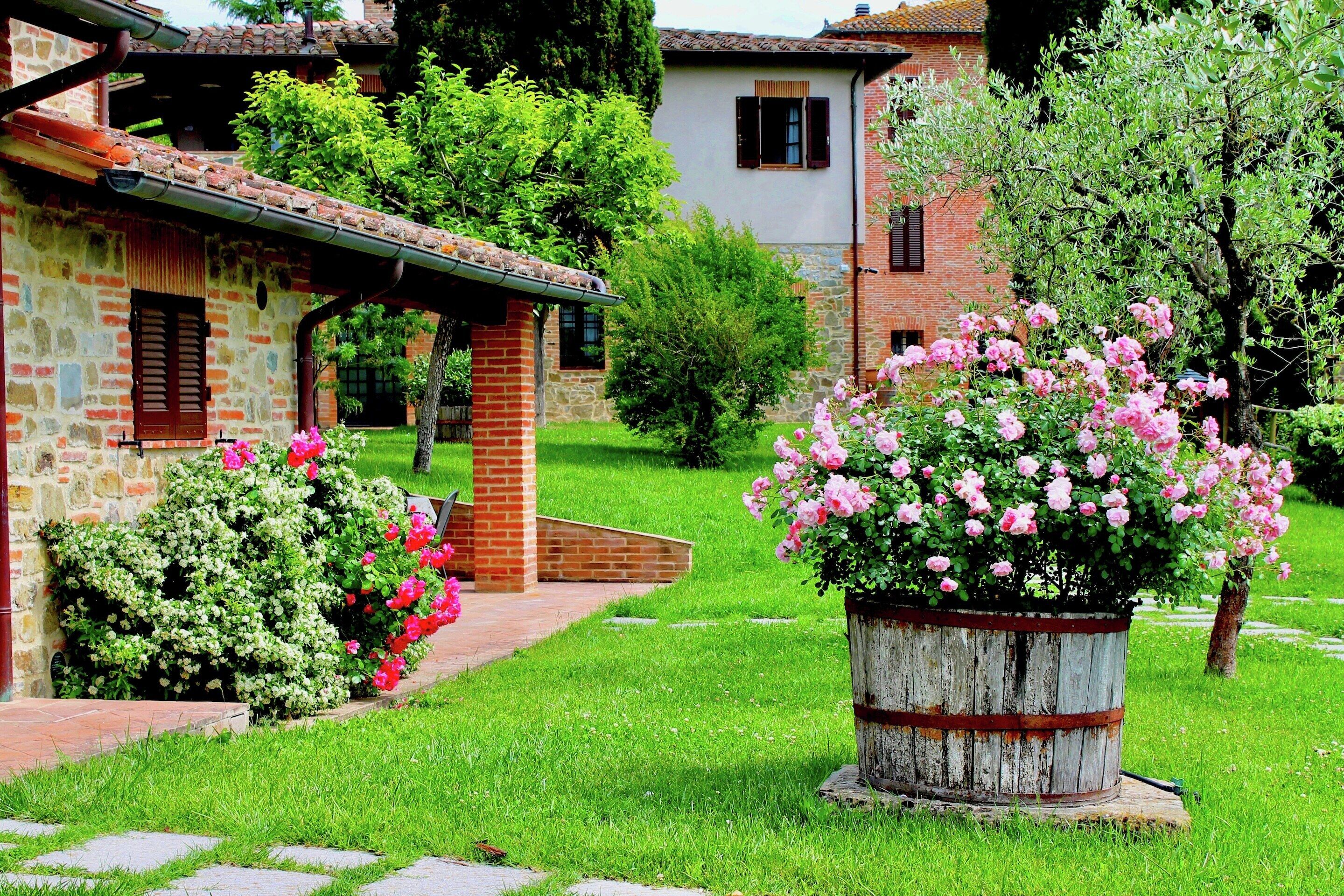 A rustic stone building with a brick chimney, a wooden barrel planter with pink flowers, and a well-maintained lawn.