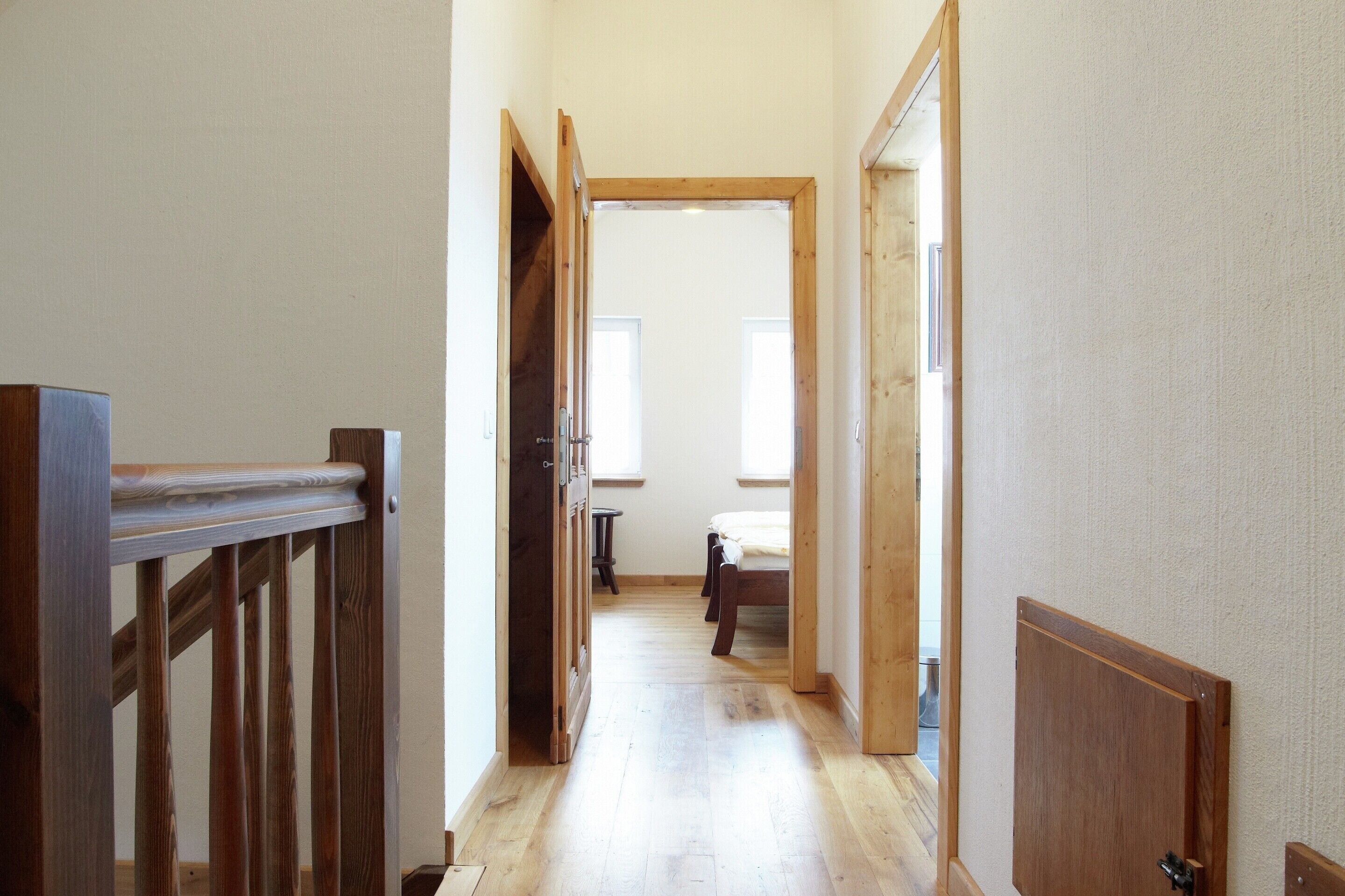 A narrow hallway with wooden flooring and doors leading to a bedroom with a bed and a window.