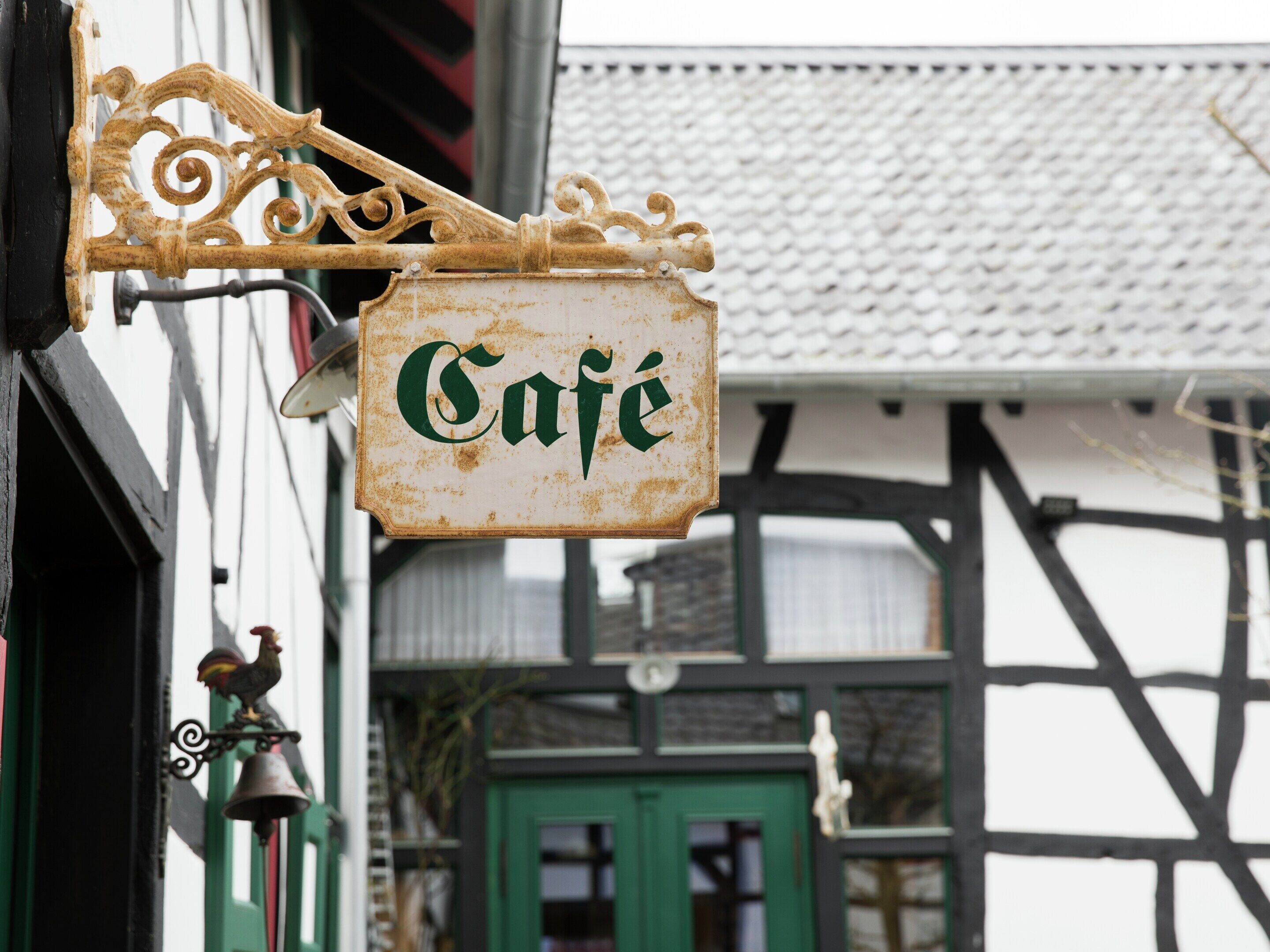 A rustic café sign with ornate metalwork, attached to a building with exposed timber framing and a green door.