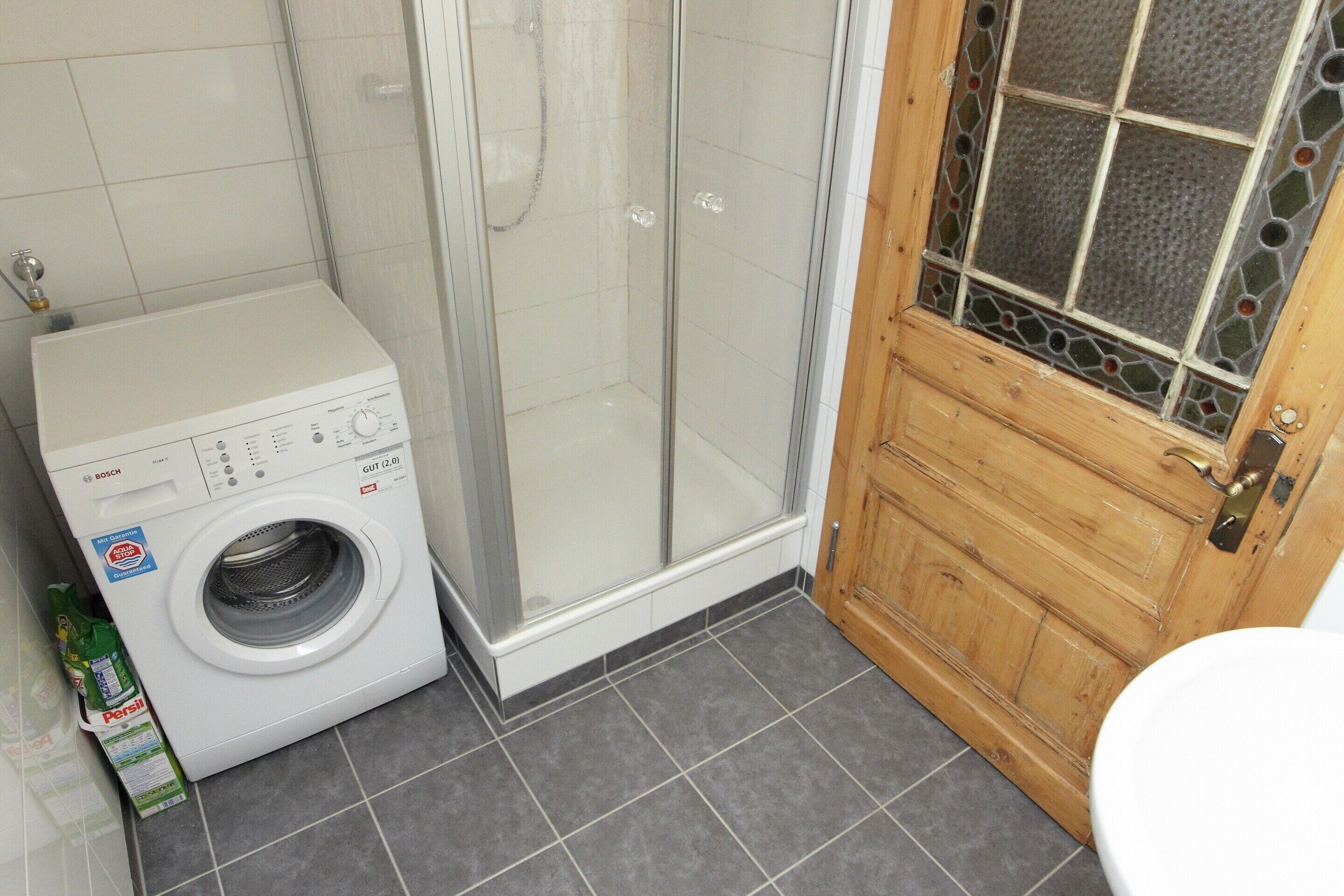 Une petite salle de bain équipée d’un lave-linge, d’un espace douche et d’une porte en bois avec un vitrage.