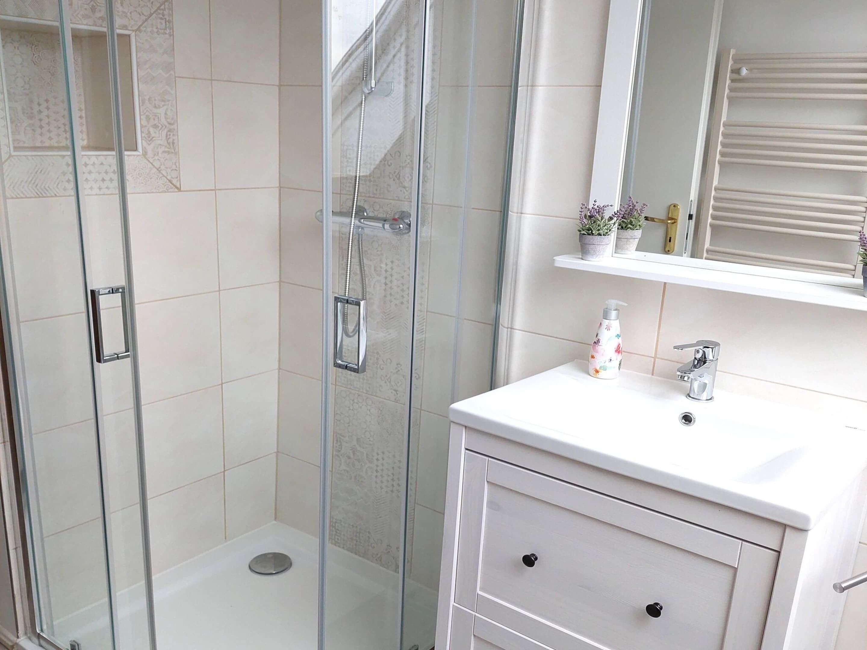 A modern bathroom with a glass-enclosed shower, white tiles, a white vanity with a mirror, and a small shelf with toiletries.