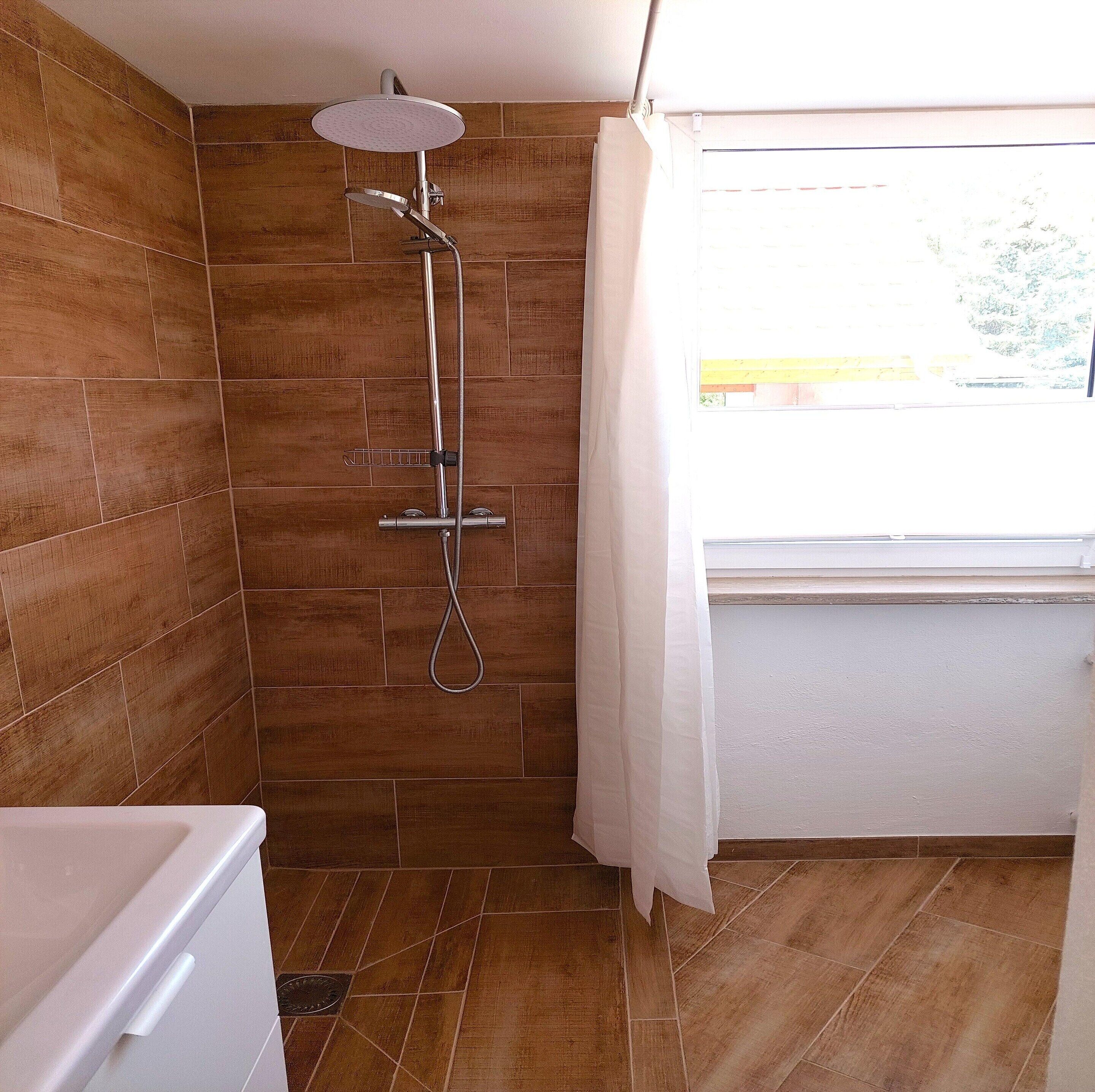 Ein Badezimmer mit Holzboden, einer Dusche inklusive Handbrause, einem Fenster mit Aussicht und einem weißen Waschbecken.
