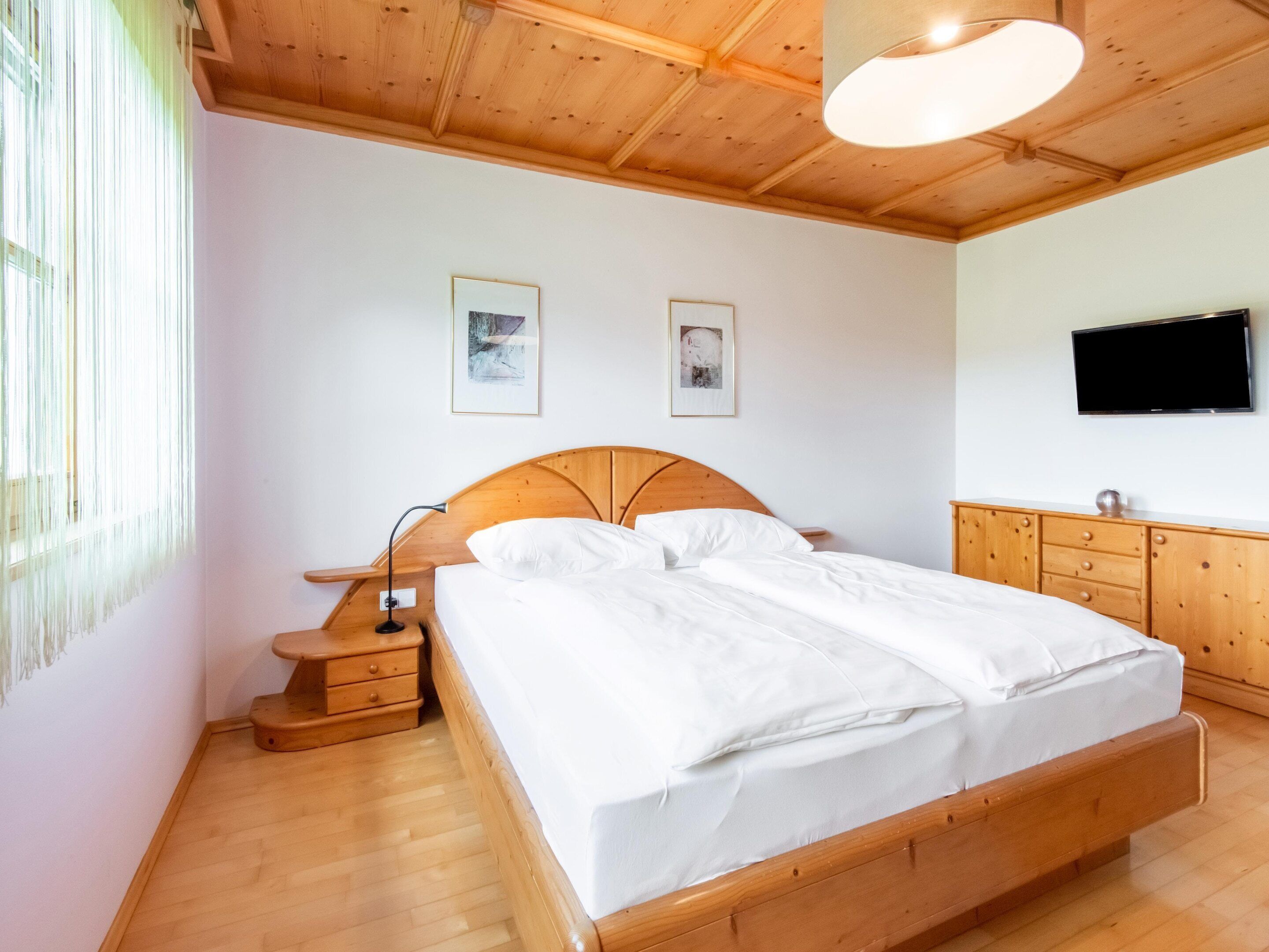 A bedroom with a wooden bed, a nightstand, a dresser, and a TV on the wall.