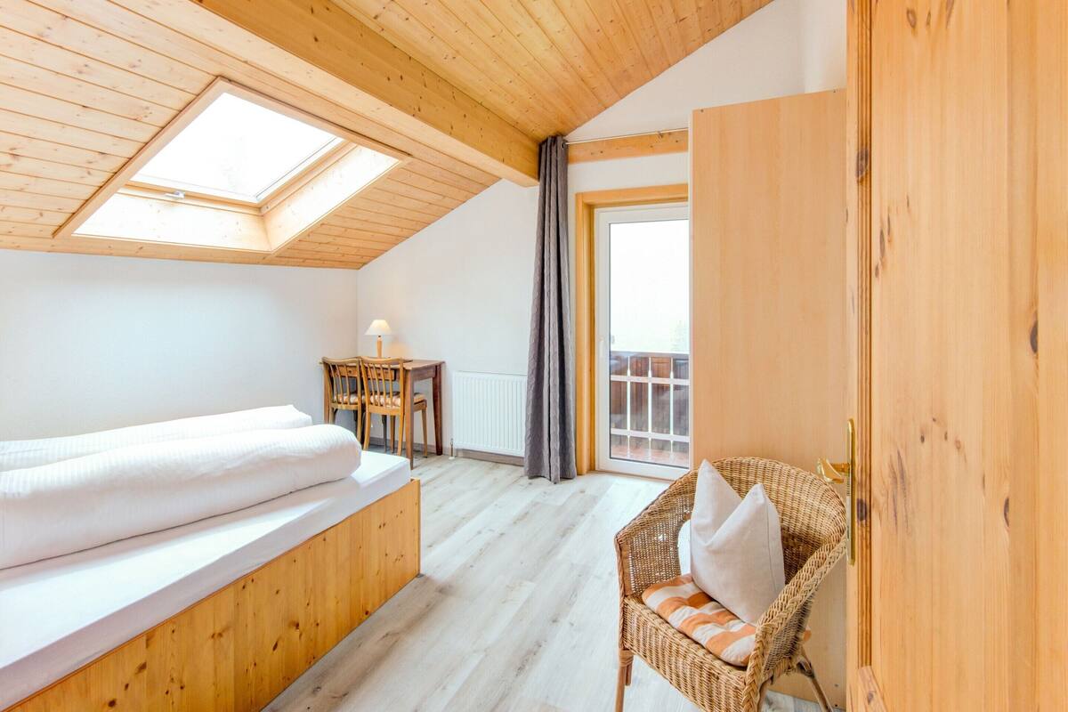 A cozy room with a wooden ceiling, a bed, a small table with a lamp, a wicker chair, and a view through a window.