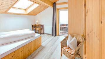 A cozy room with a wooden ceiling, a bed, a small table with a lamp, a wicker chair, and a view through a window.