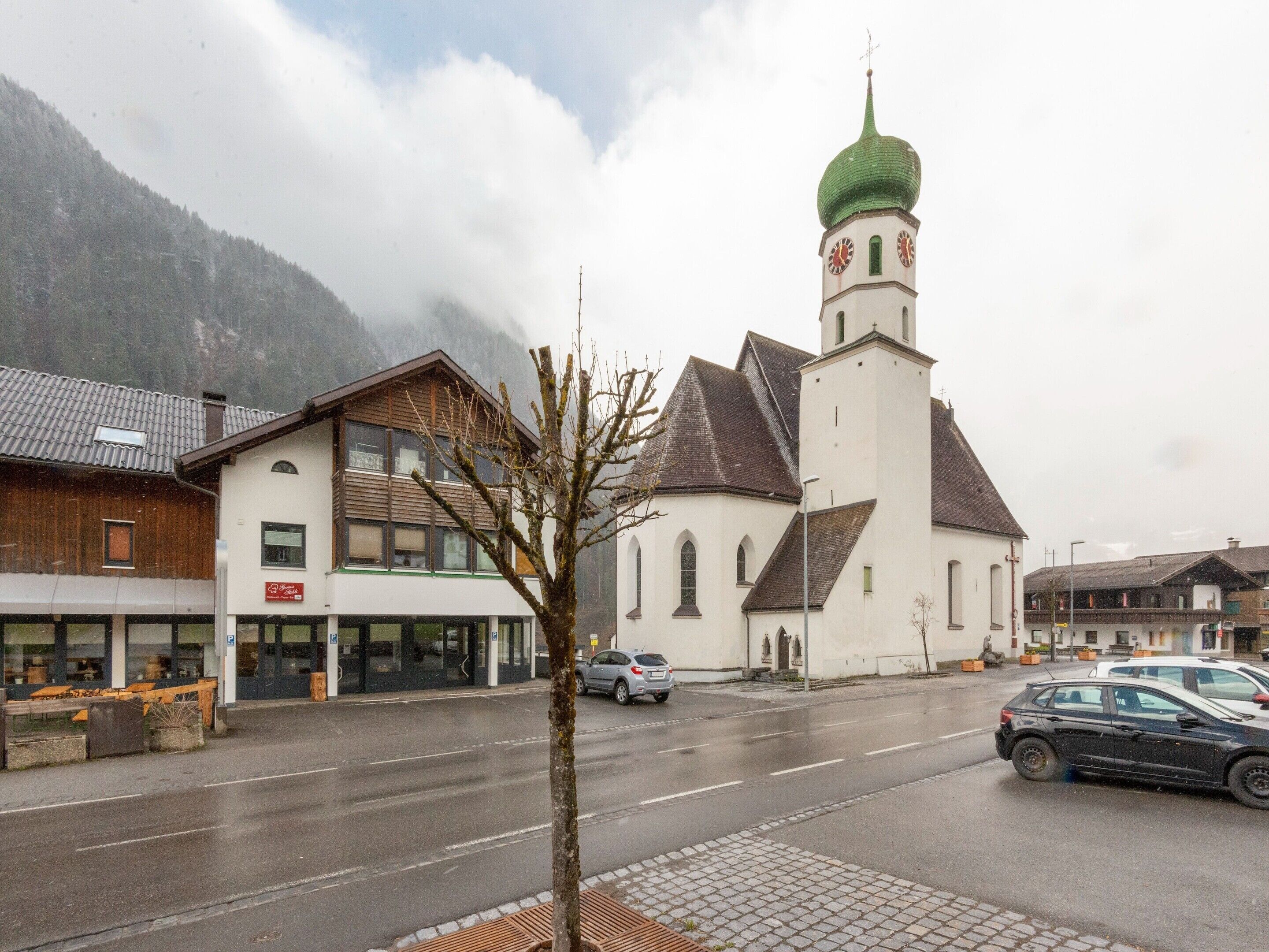 Una iglesia con una cúpula verde y una torre del reloj, un edificio con un letrero, y un árbol en primer plano.