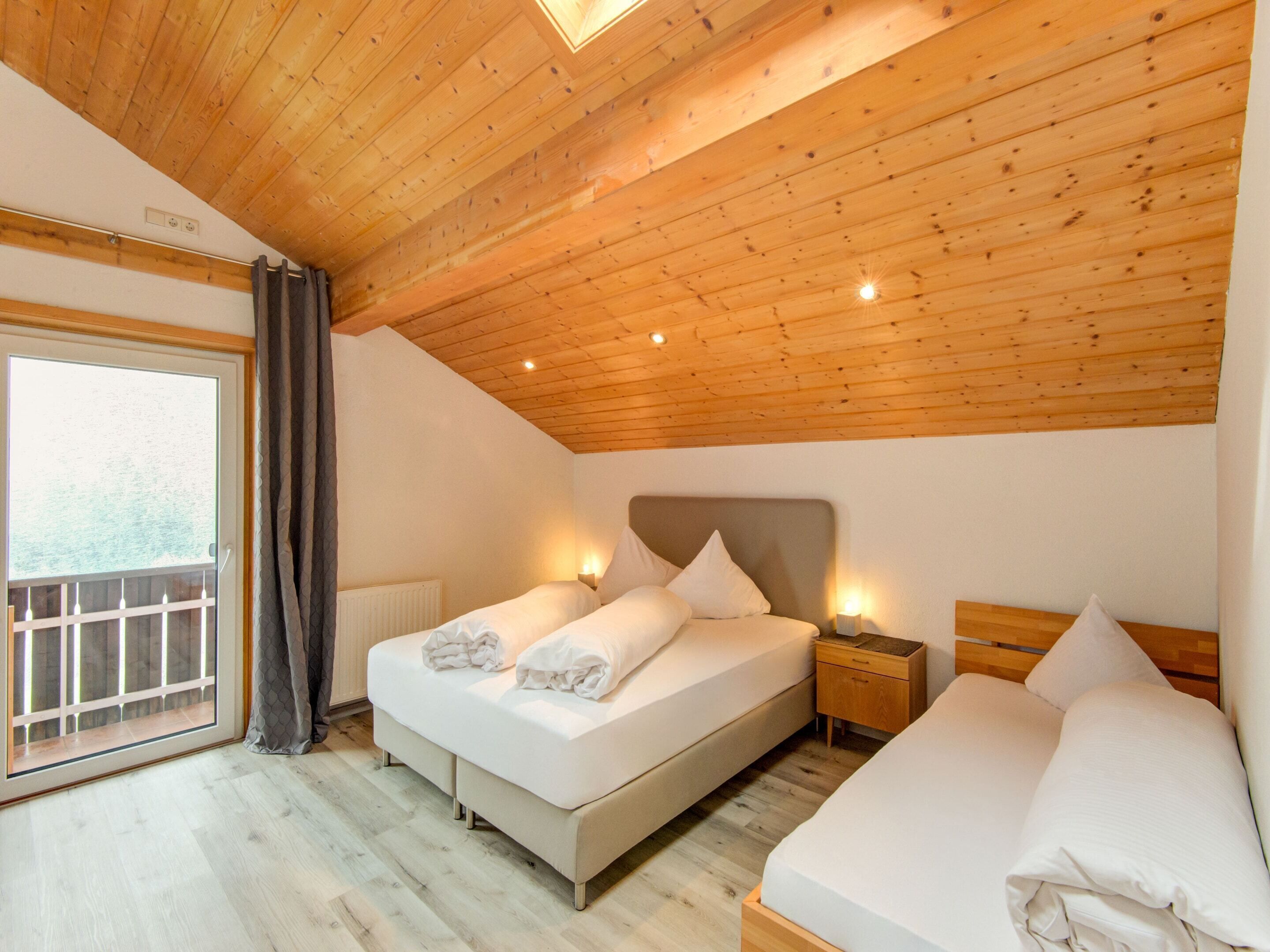 A bedroom with a wooden ceiling, a large window with curtains, a bed with pillows, and a bedside table with a lamp.