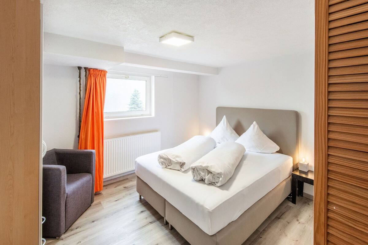 A bedroom with a bed, a chair, a window with curtains, and a radiator.
