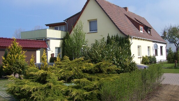 Une maison à deux étages, dotée d’un toit en pente, d’un balcon et d’un jardin bien entretenu.