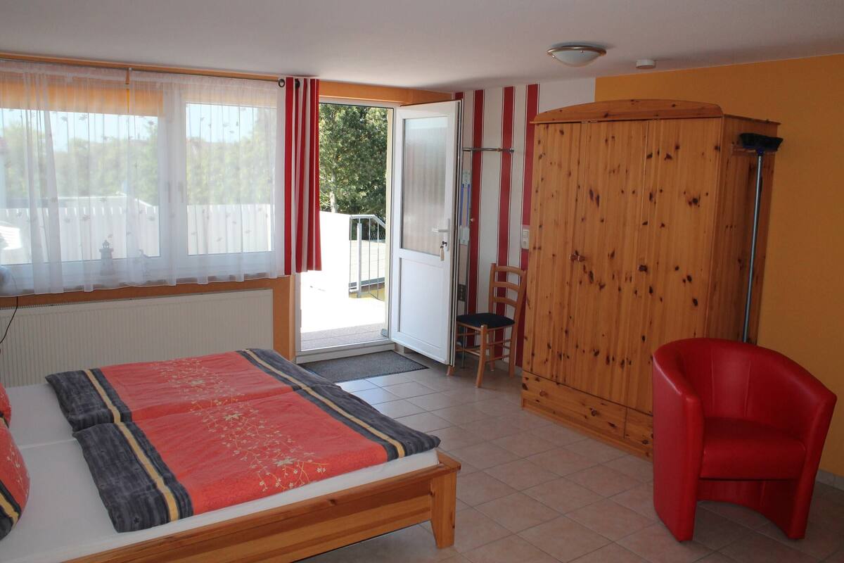 A bedroom with a bed, a red chair, a wooden wardrobe, and a sliding glass door leading to a balcony.