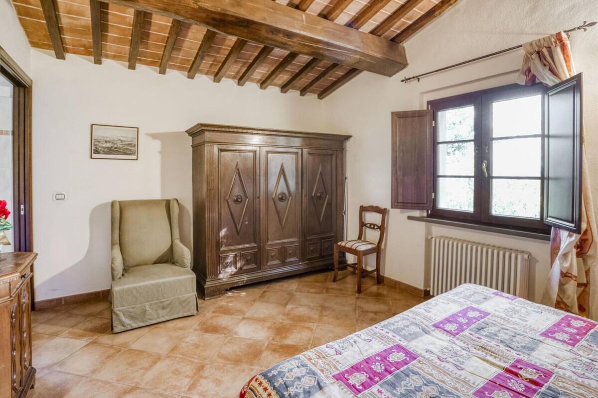 A bedroom with a wooden ceiling, a large wardrobe, a bed with a patterned bedspread, a chair, and a window with curtains.
