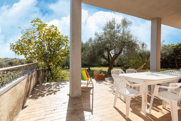 Un patio techado al aire libre con una mesa y sillas blancas, un caballete pequeño y vistas a árboles y un área con césped.