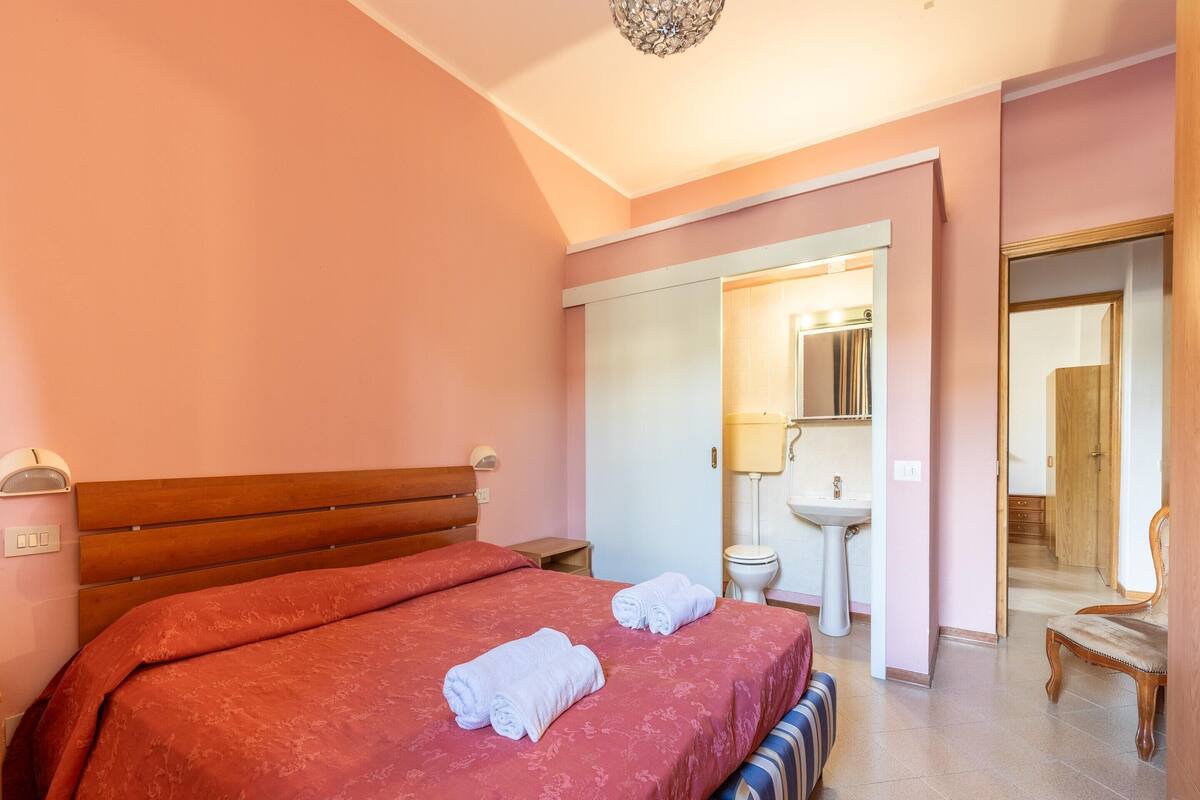 Ein Schlafzimmer mit einem großen Bett, einem Nachttisch, einem Waschtisch, einem Badezimmer mit Waschbecken und Toilette sowie einem Spiegel.