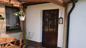 A wooden bench outside a building with a door and windows, a hanging flower pot, and a lantern on the porch. - Beautiful Apartment in Robertsdorf With Garden (Blowatz)