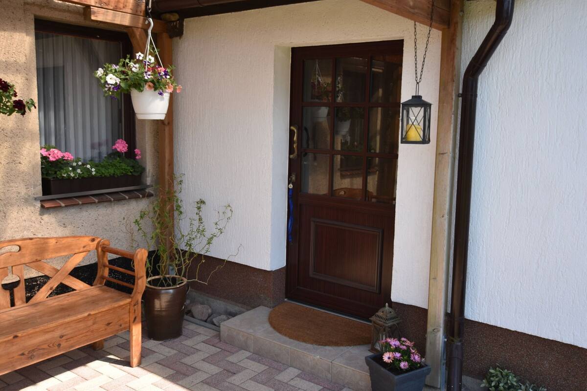 A wooden bench outside a building with a door and windows, a hanging flower pot, and a lantern on the porch.