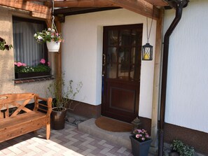A wooden bench outside a building with a door and windows, a hanging flower pot, and a lantern on the porch. - Beautiful Apartment in Robertsdorf With Garden (Blowatz)