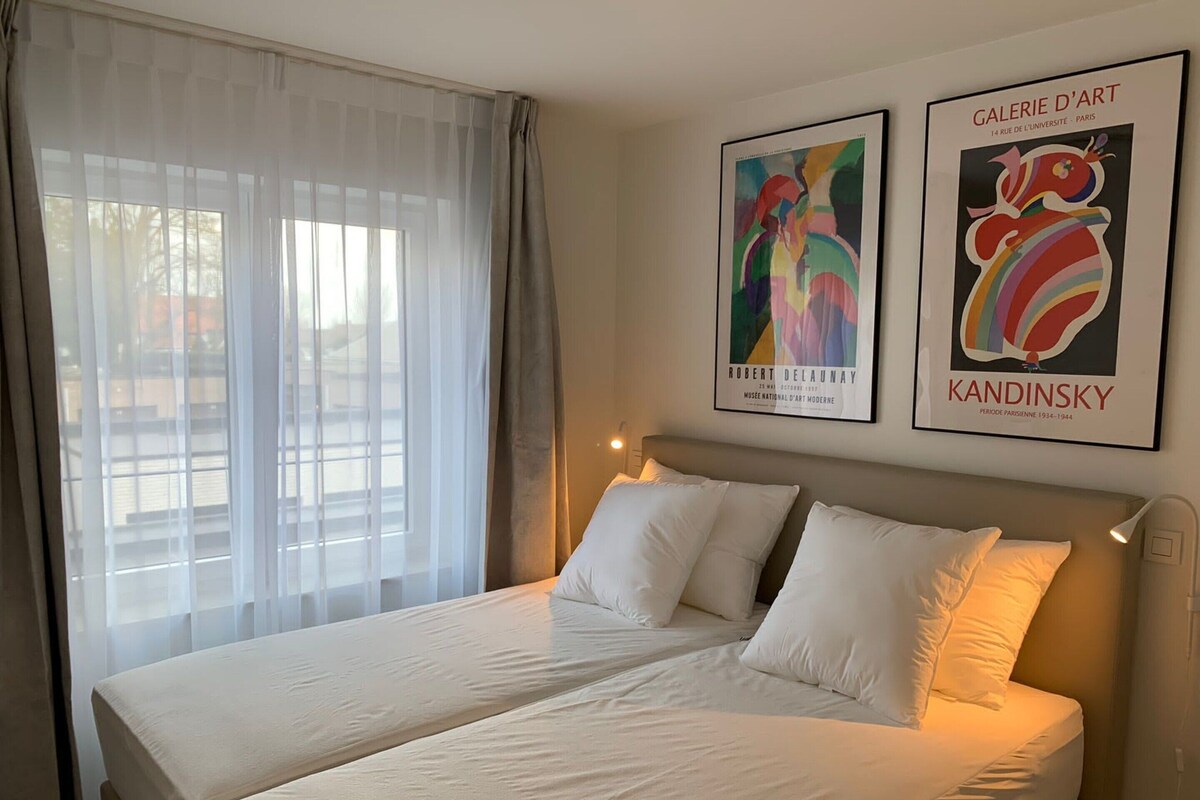 A hotel room with a bed, white curtains, and two framed artworks on the wall.