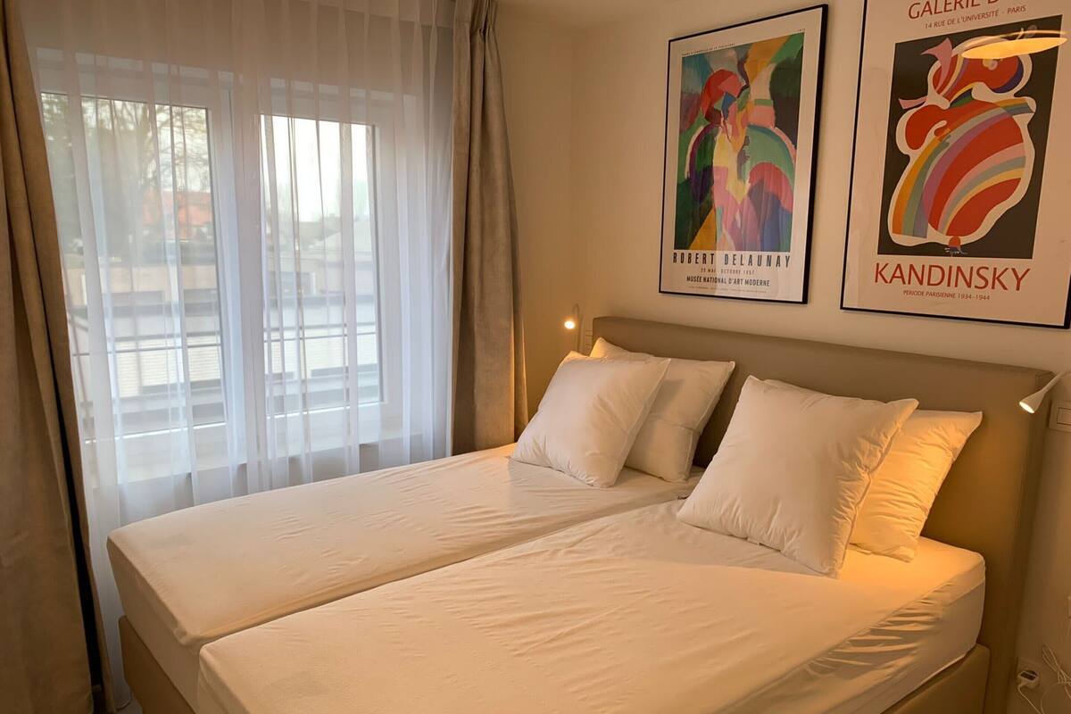 A double bed with white linens, a headboard, and a window with sheer curtains.