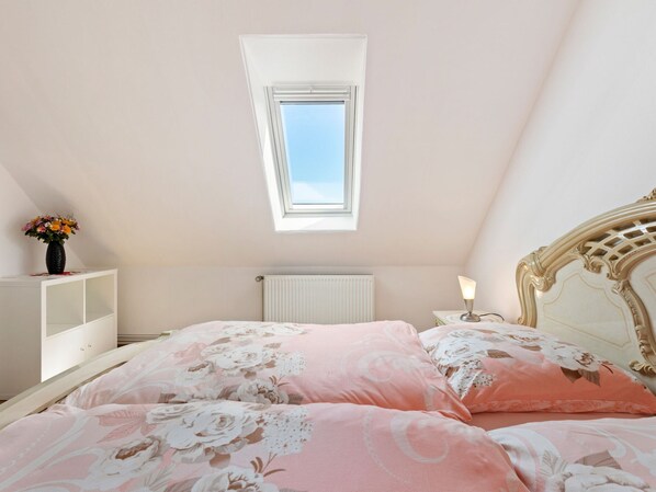Ein Schlafzimmer mit schrägen Wänden, einem Oberlicht, einem weißen Heizkörper, einem Nachttisch mit Lampe und einem Bett mit floralem Bettbezug.