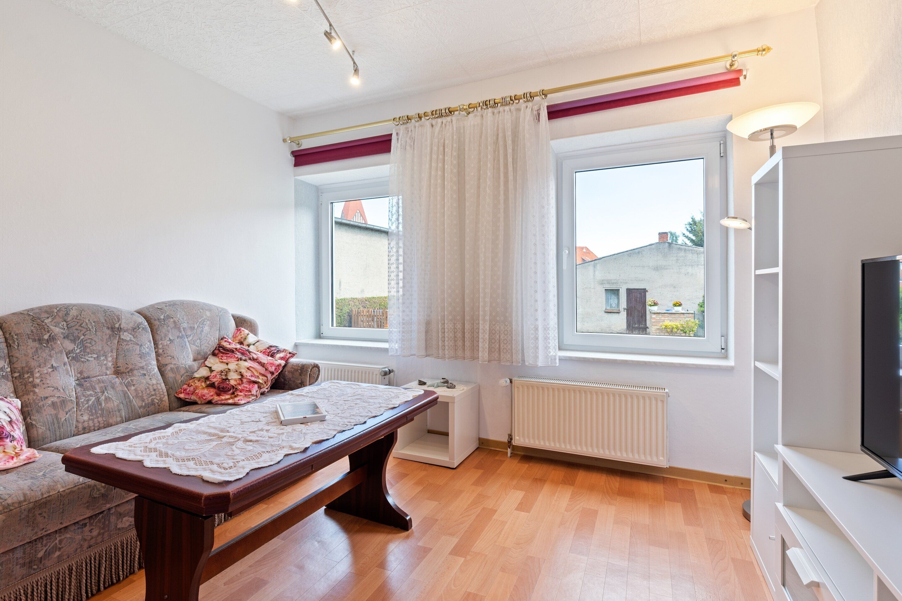 Ein Wohnzimmer mit einer Couch, einem Tisch mit Spitzenbettwäsche, einem Fernseher und zwei Fenstern mit Vorhängen.