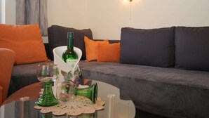 A living room with a dark grey sofa, orange cushions, a glass coffee table with a green vase and a glass pitcher, and a wall-mounted light fixture. - Modern Apartment With Garden Near the Petzen ski Area in Eberndorf / Carinthia (Eberndorf)