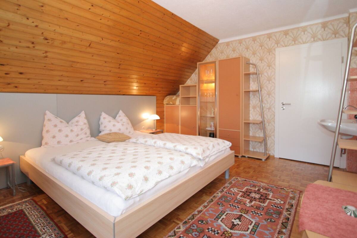 A bedroom with a wooden ceiling, a bed with patterned bedding, a wardrobe, and a bathroom with a sink.