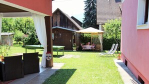 A covered patio area with a table and chairs, a grassy lawn, and a wooden building in the background. - Modern Apartment With Garden Near the Petzen ski Area in Eberndorf / Carinthia (Eberndorf)