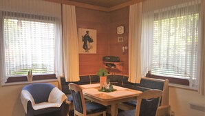 A dining area with a wooden table, blue upholstered chairs, and a bench. There are two windows with blinds, a clock, and a small cactus plant on the windowsill. - Modern Apartment With Garden Near the Petzen ski Area in Eberndorf / Carinthia (Eberndorf)