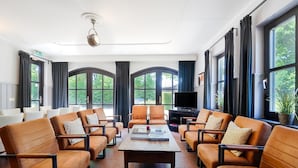 Un salón amplio con sofás de cuero dispuestos alrededor de una mesa de centro, ventanas grandes con cortinas y un ventilador de techo.