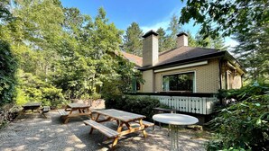 Ein rustikales Blockhaus mit Backsteinfassade, umgeben von dichtem Wald. Auf dem Grundstück befinden sich Picknicktische und ein kleiner runder Tisch.