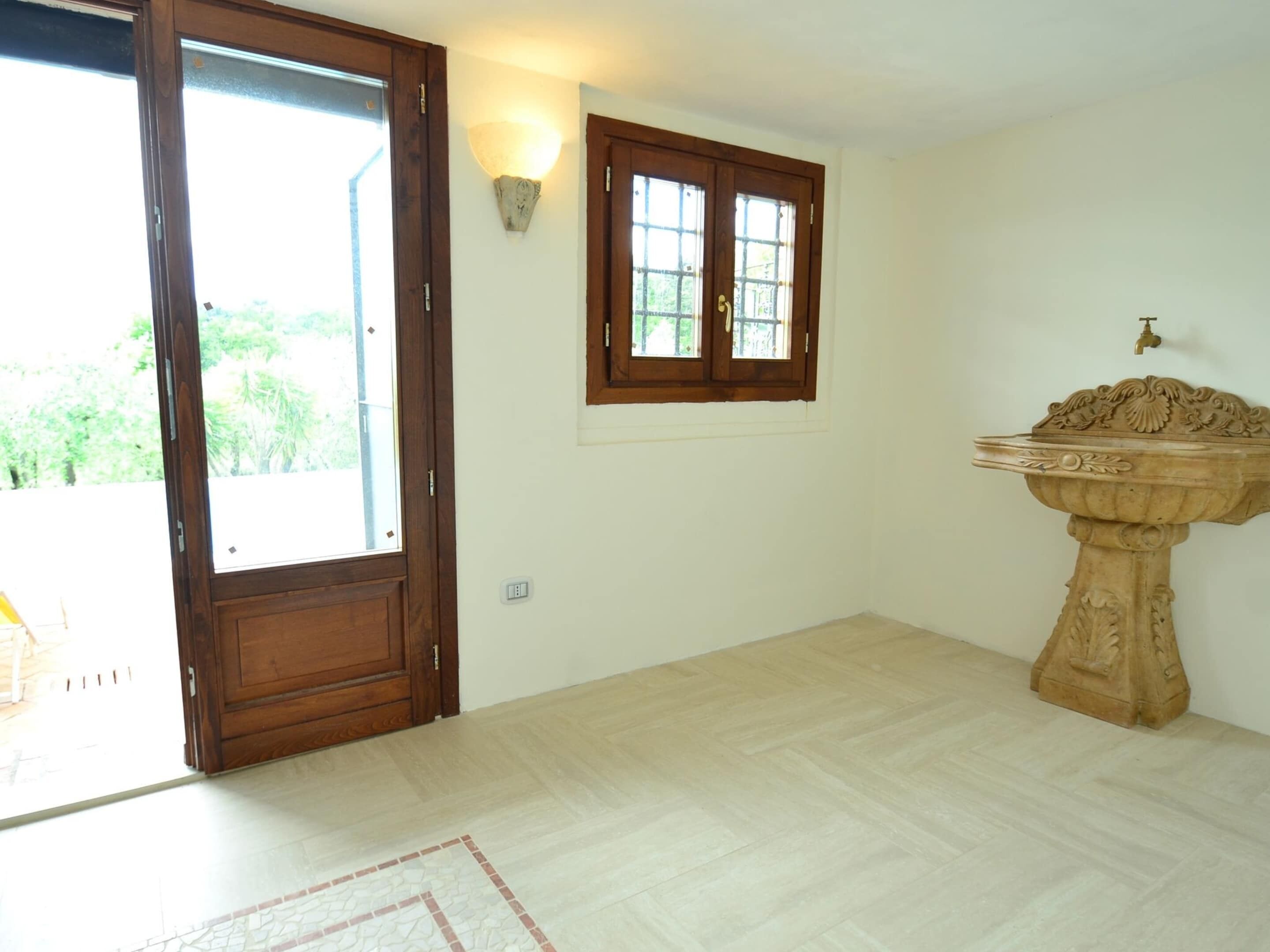 A room with a wooden door, a window with a view, and a stone pedestal with a basin.