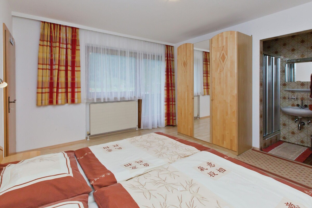 Habitación de hotel con cama, ventana con cortinas traslúcidas, armario de madera, baño con lavamanos y espejo, y radiador.