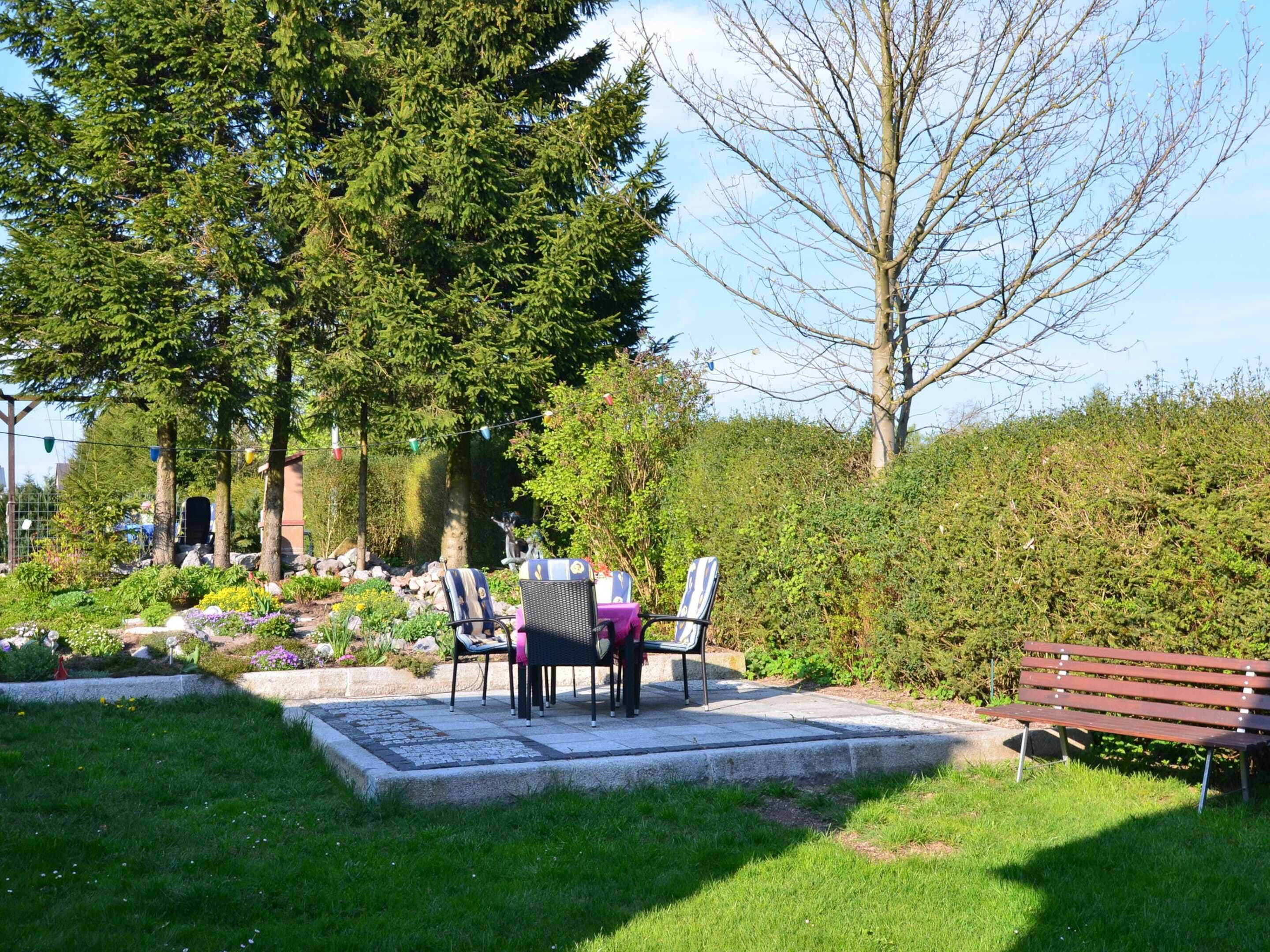 Ein Sitzbereich im Freien mit Tisch und Stühlen, umgeben von Pflanzen und einer Hecke.