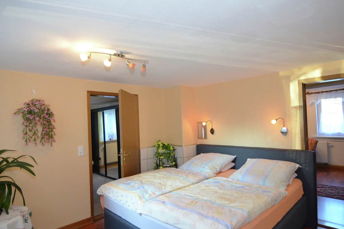 A bedroom with a double bed, a potted plant, a hanging flower arrangement, and a window with curtains.