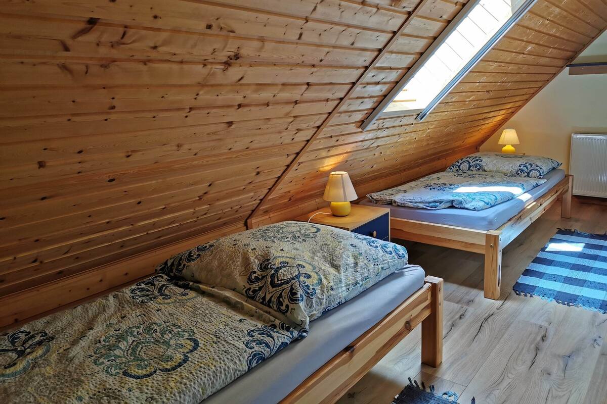 A cozy attic bedroom with wooden beams, two beds with patterned bedding, a small table with a lamp, and a view through a skylight.